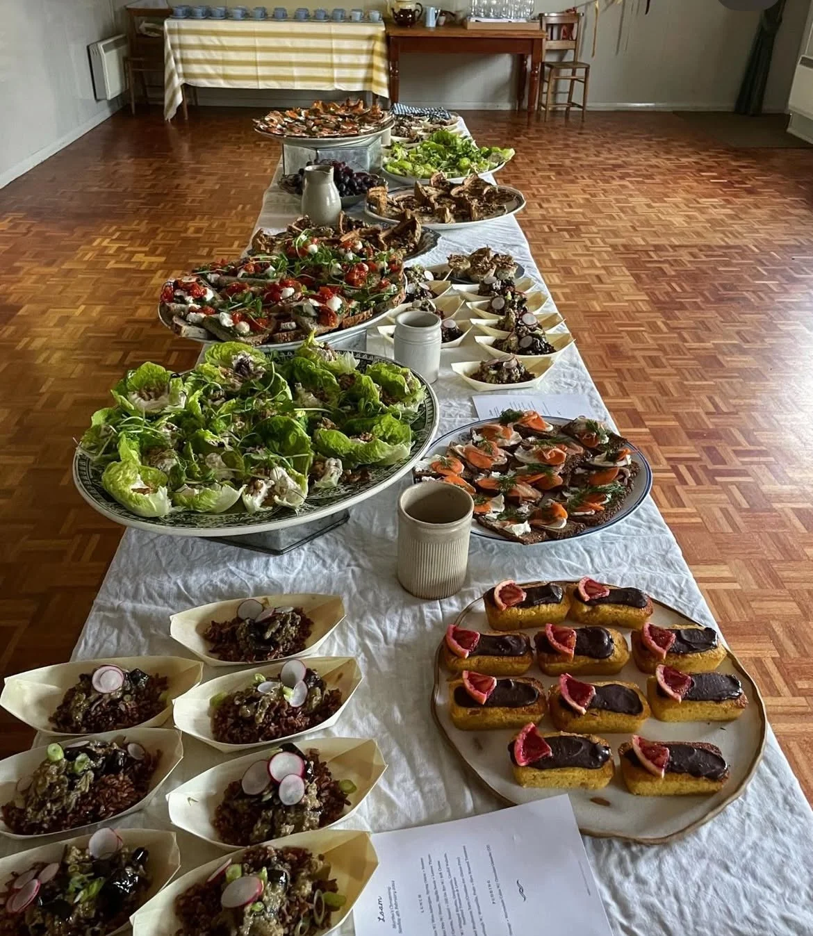 A long table covered with a white tablecloth displays various dishes including salads, appetizers, and desserts, with a wooden floor and a background of a room with a table and chairs.