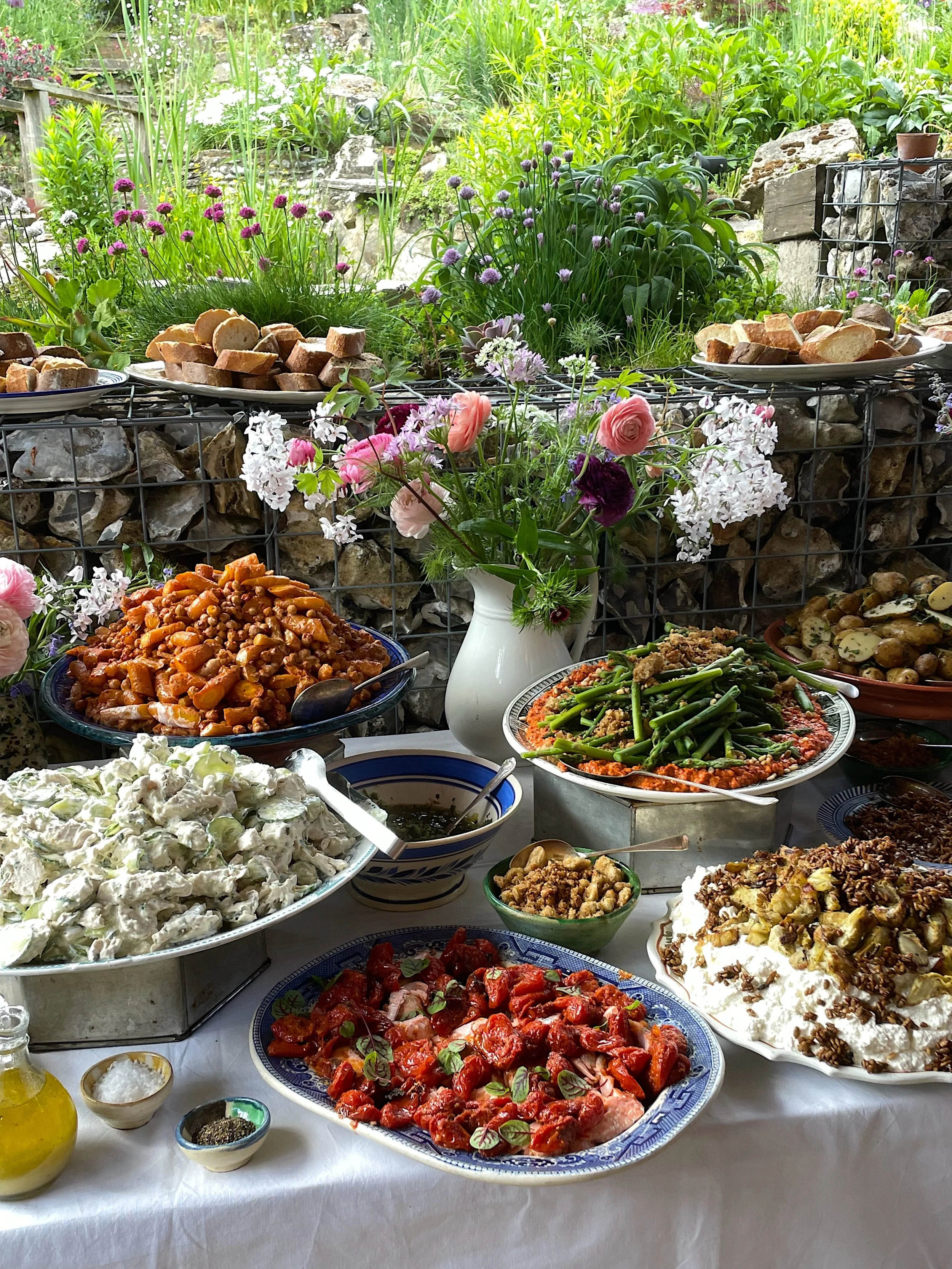 A table filled with various dishes of salad, cooked vegetables, and bread, with a beautiful floral arrangement in a vase, set outdoors in a garden with greenery and flowers in the background.