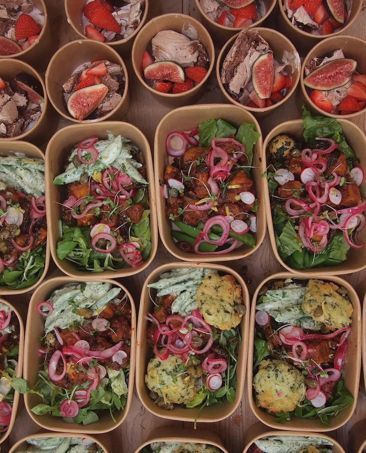 Multiple small bowls of salad with vegetables and herbs, alongside bowls of sliced strawberries and figs.