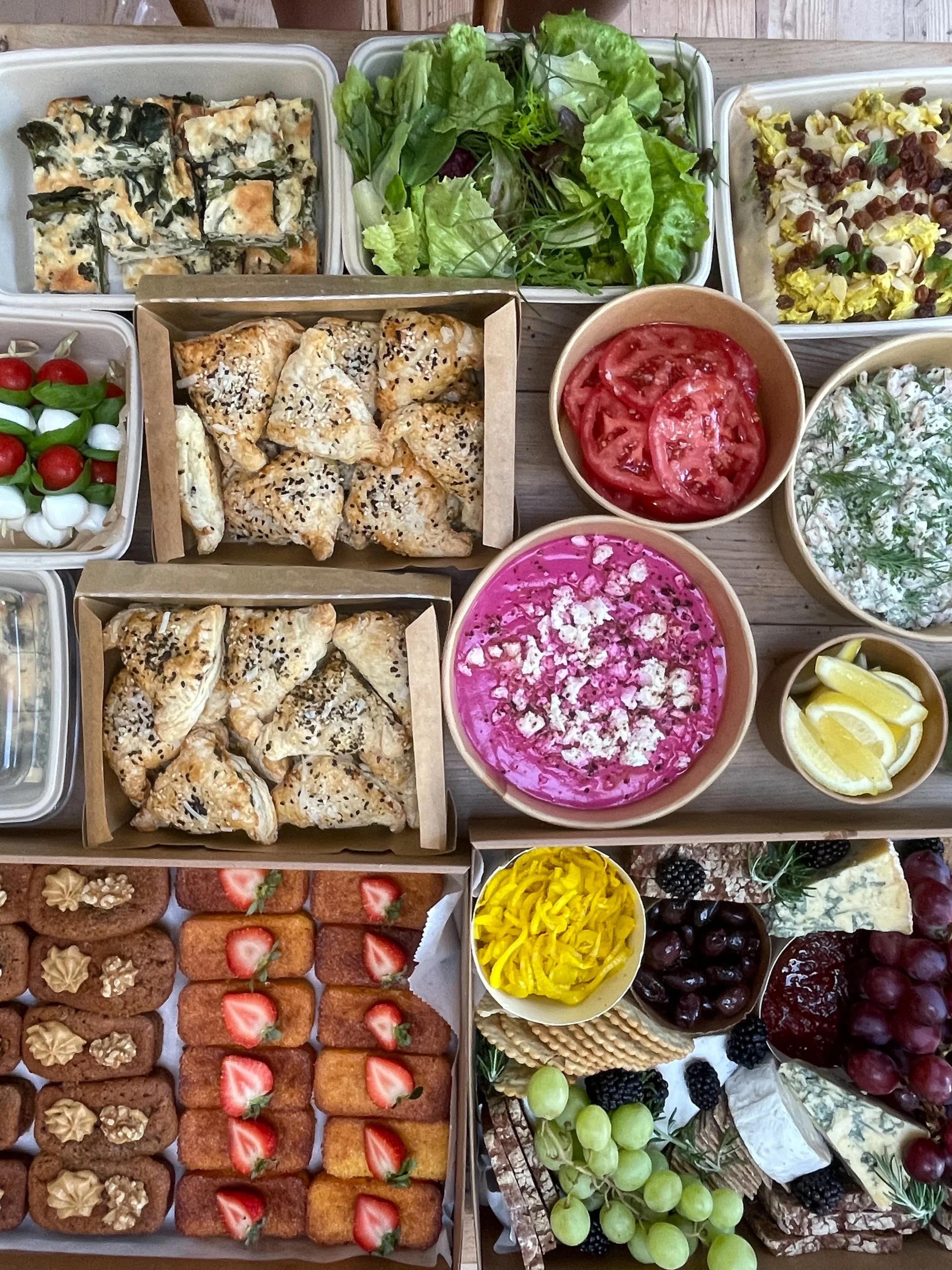 Assorted appetizers and desserts on a table, including spinach and cheese squares, salad greens, a tray of small tomatoes, scones topped with black sesame seeds, a bowl of sliced tomatoes, a bowl of pink beet dip with feta cheese, a bowl of creamy he