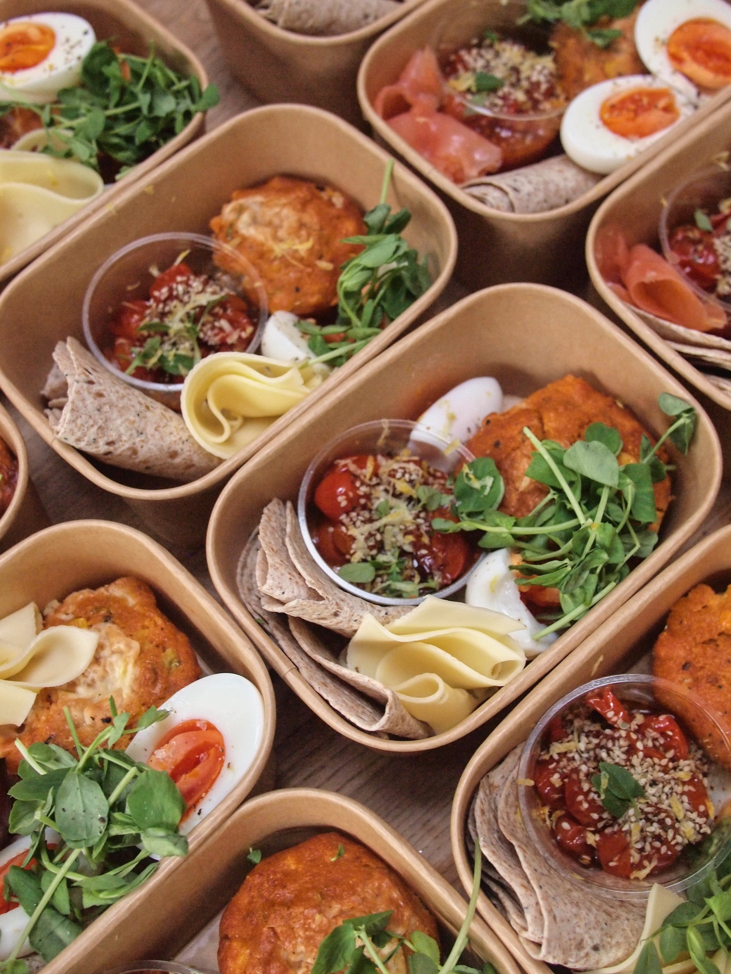Multiple bento boxes containing soft-boiled eggs, roasted tomatoes, microgreens, flatbread, and a baked item.