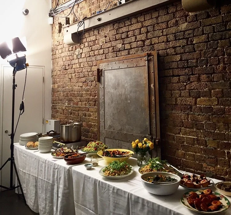 Buffet table set against a brick wall with various bowls of salad and dishes, plates stacked on the side, a vase with yellow flowers, and a lighting stand nearby.