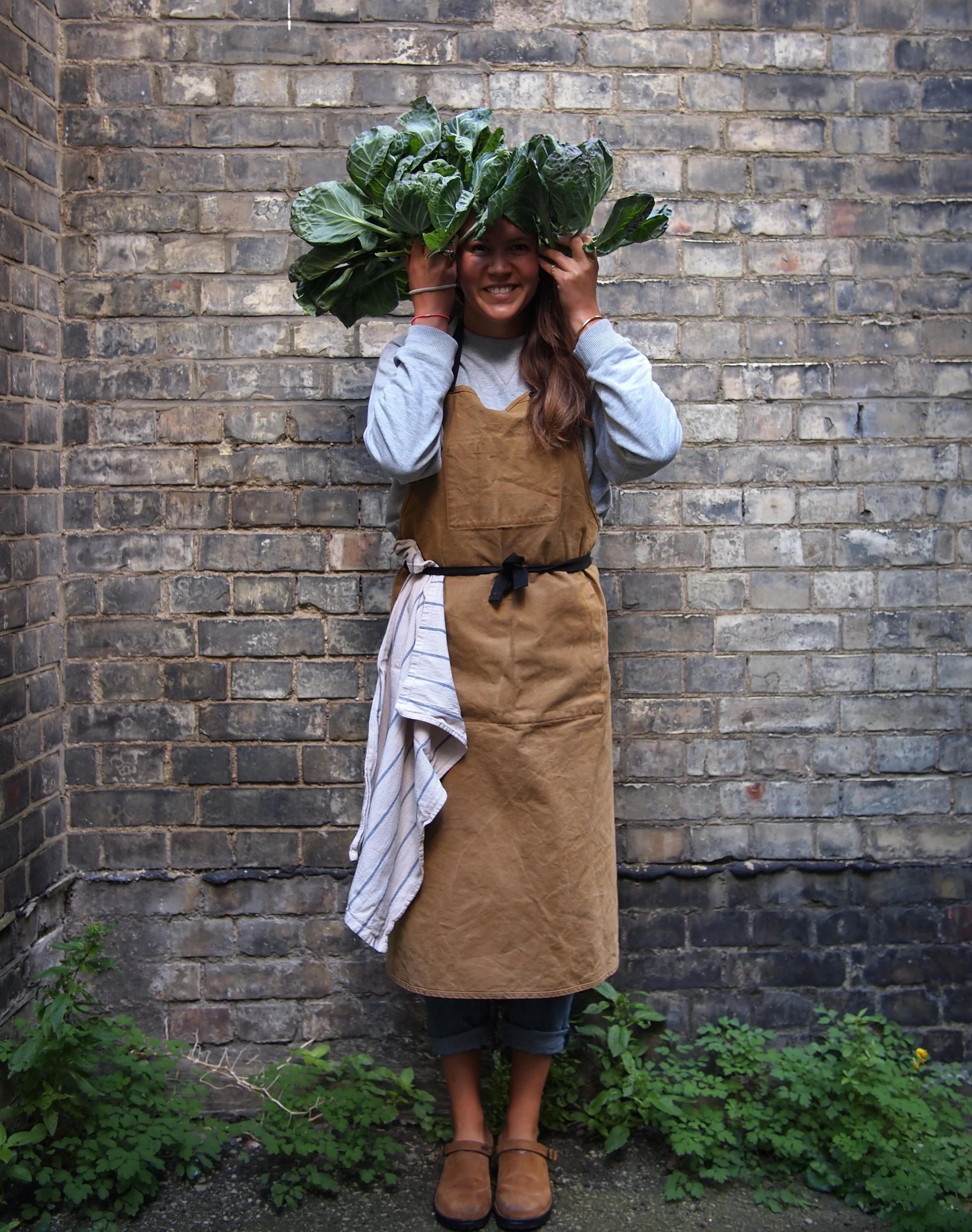A woman standing outdoors against a brick wall, smiling, carrying a large bunch of leafy greens on her head, wearing a brown apron, blue sweatshirt, dark pants, and tan shoes.