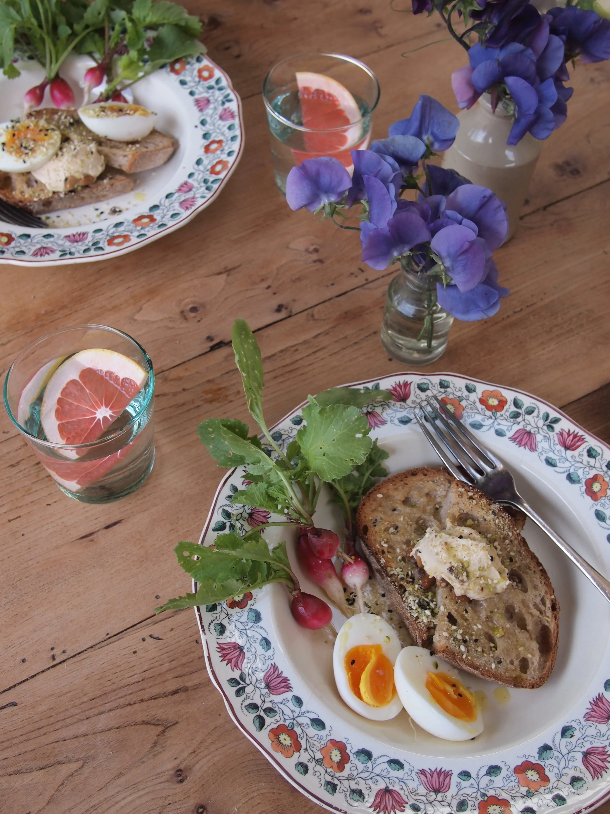 A breakfast with toast topped with butter and seeds, soft-boiled eggs, radishes with greens, and glasses of grapefruit water, set on a wooden table with purple flowers in vases.