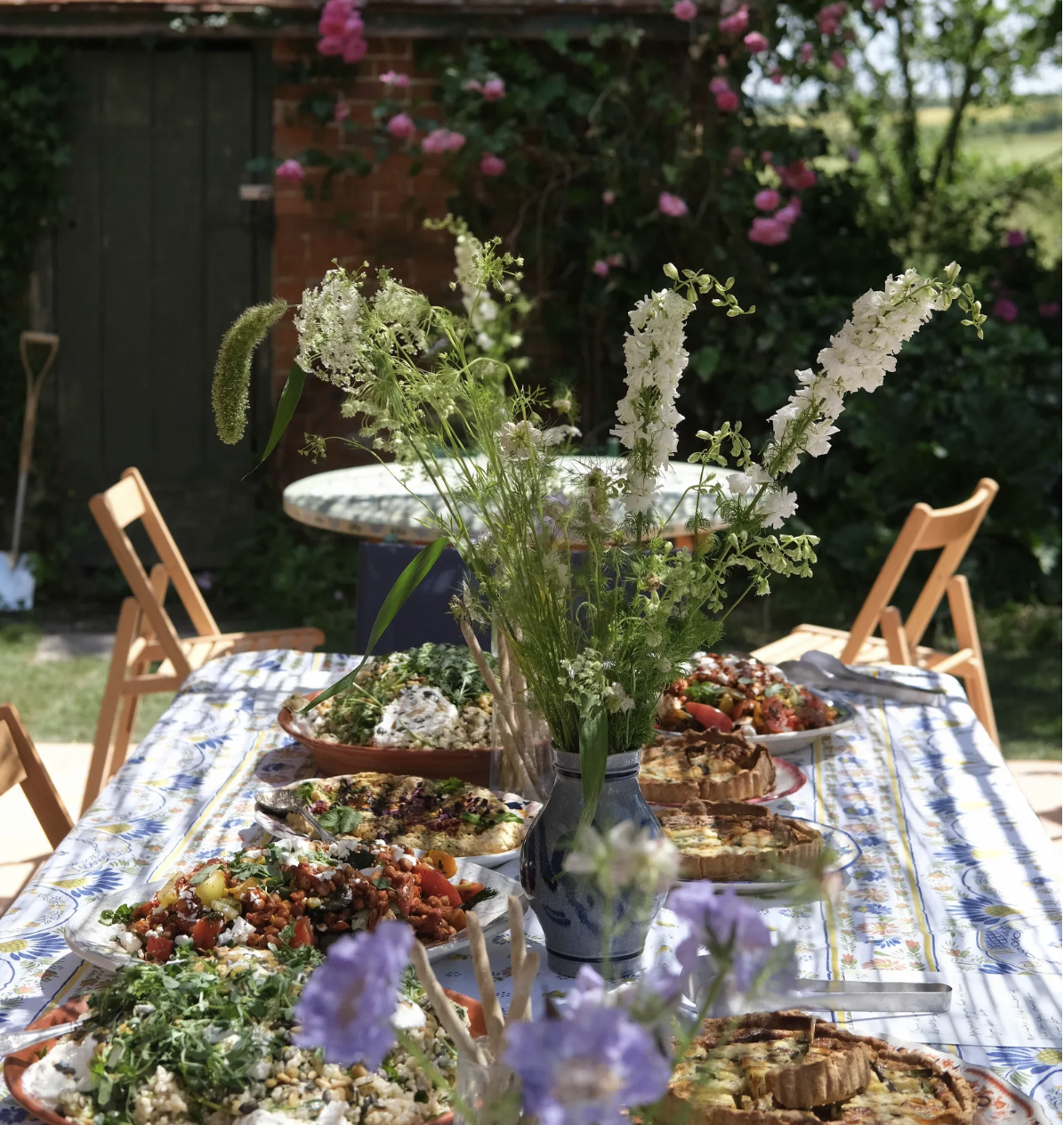 An outdoor table set with various dishes and a vase of white and green flowers, surrounded by garden chairs, with a background of greenery and pink flowers.