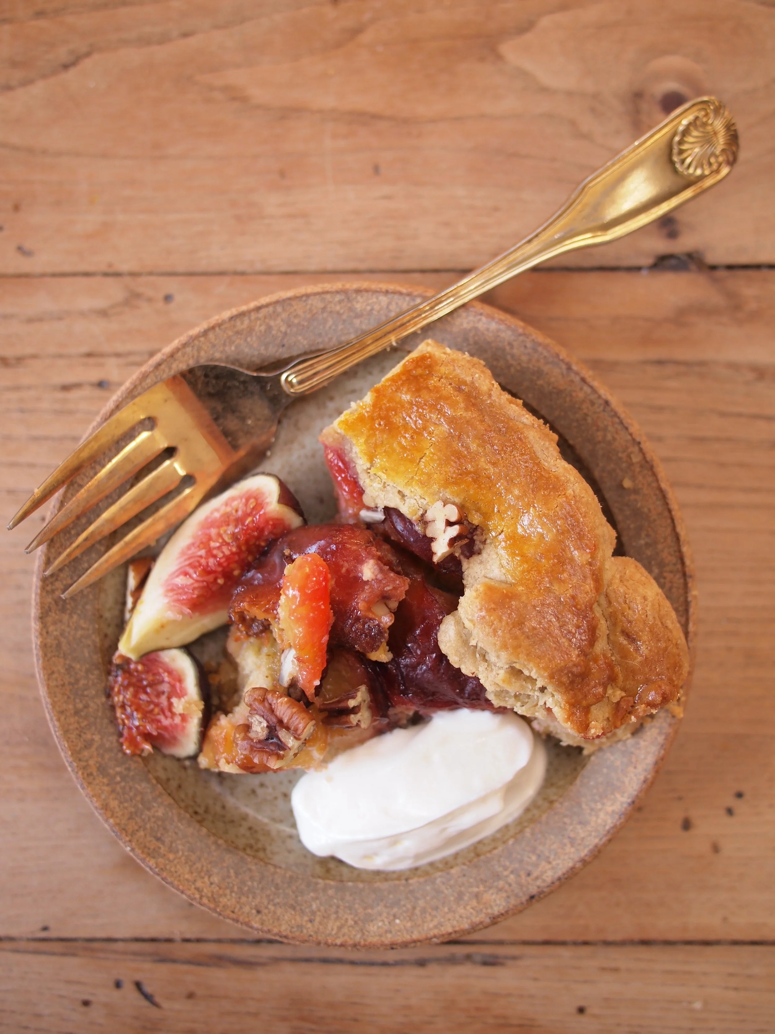 A bowl of peach cobbler with sliced figs, chopped pecans, a dollop of whipped cream, and a gold fork on a wooden table.