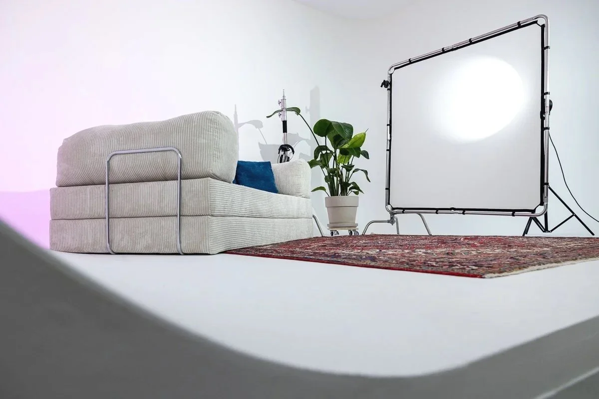 Ein Wohnzimmer-Setup mit einem weißen Sofa, einer roten Teppich, einer grünen Pflanze in einem weißen Topf, einer Studiolampe und einem Beleuchtungsgerät auf einer weißen Wand.