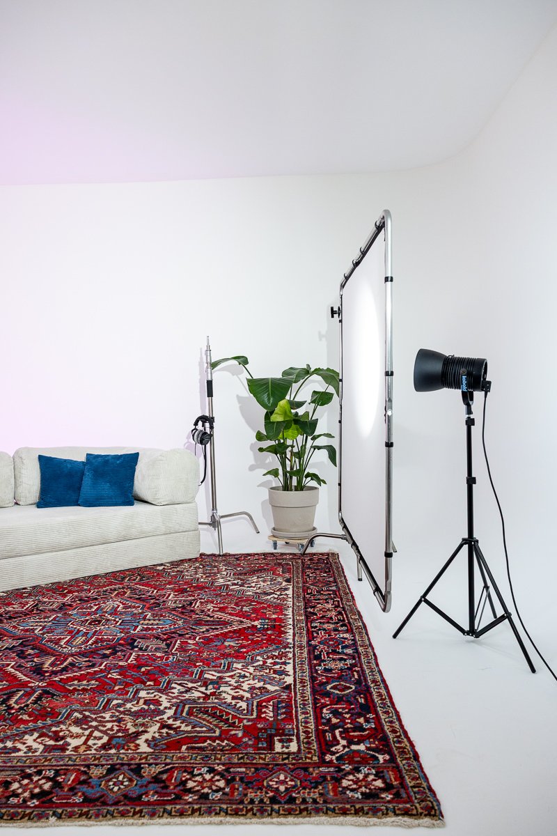 Ein Studio mit weißer Wand, einem roten orientalischen Teppich, einem weißen Sofa mit blauen Kissen, einer grünen Zimmerpflanze, einem Fototuch, einer Studiolichtquelle und einer Fotowand