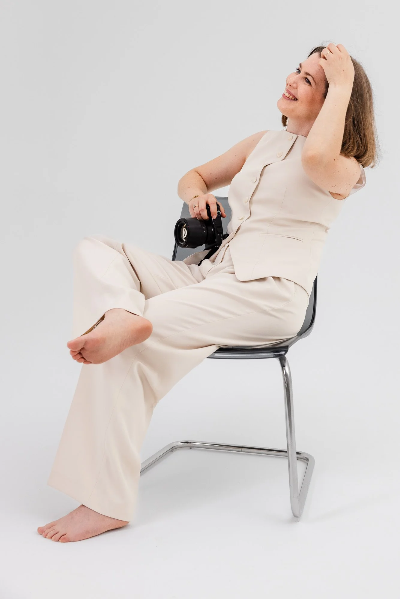 Eine lachende Frau in einem weißen Outfit sitzt auf einem Stuhl, hält eine Kamera und streicht sich durch die Haare, vor einem weißen Hintergrund.