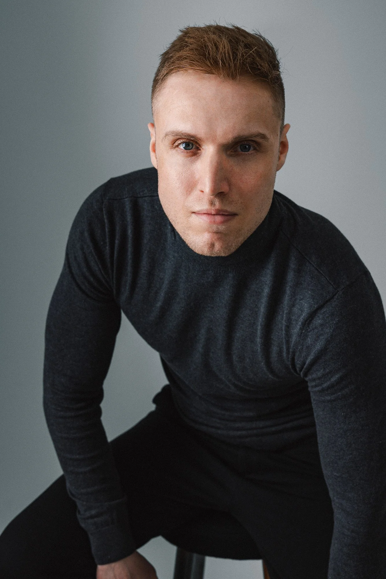 Ein junger Mann mit kurzen braunen Haaren und blauen Augen sitzt auf einem Stuhl vor einem grauen Hintergrund, trägt einen schwarzen Pullover und blickt direkt in die Kamera.