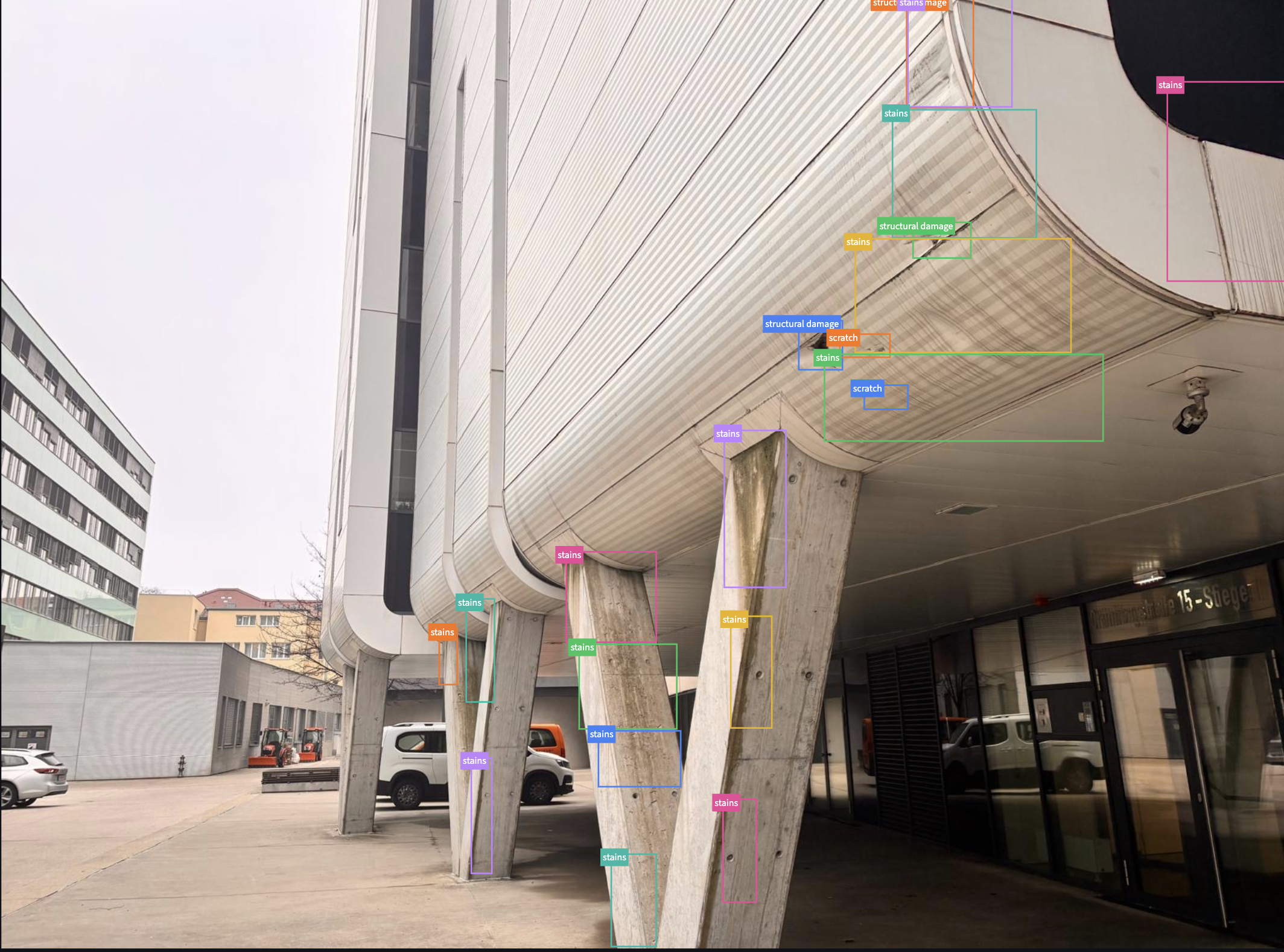 Eine moderne, architektonisch gestaltete Gebäudefassade mit auffälligen, schrägen Betonsäulen und einer geschwungenen Dachkonstruktion. Es gibt mehrere Fahrzeuge und eine Parkfläche im Vordergrund. Die Fassade wird durch eine Vielzahl farblich gekennzeichneter Linien und Labels für Schäden wie Risse, Abplatzungen, Flecken und Staub markiert.