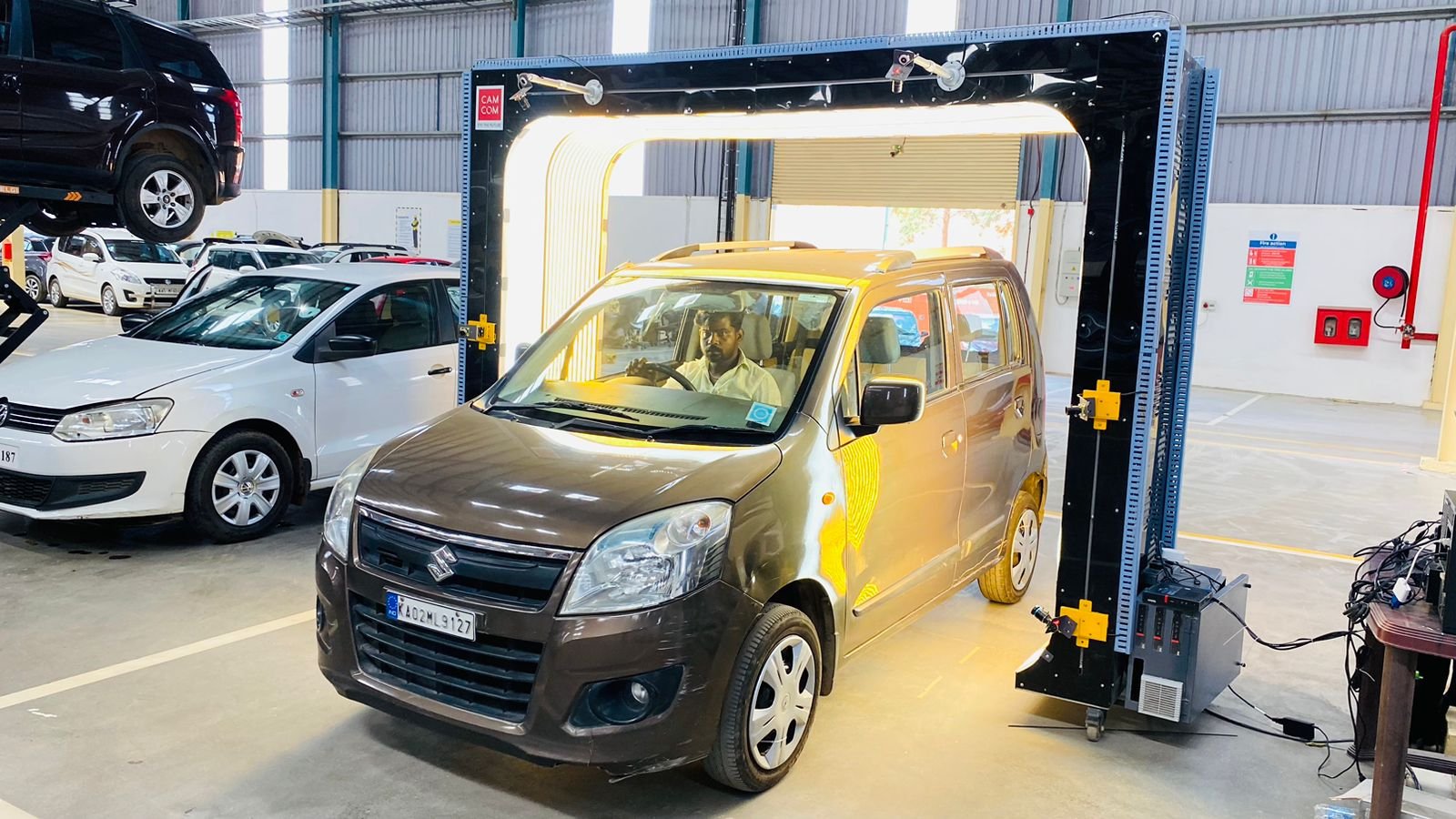 A brown Suzuki car inside a vehicle testing chamber with a technician sitting in the driver’s seat, surrounded by other parked cars in a spacious indoor lot.