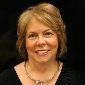 Smiling woman with short blonde hair and a necklace, wearing a black top, on a dark background.