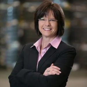 Person wearing a black suit with pink shirt, smiling with arms crossed, blurred background.