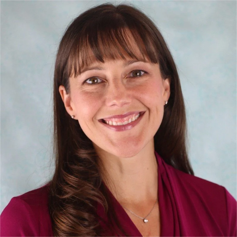 Smiling woman with long hair and bangs, wearing a maroon top and a delicate necklace, against a light blue background.