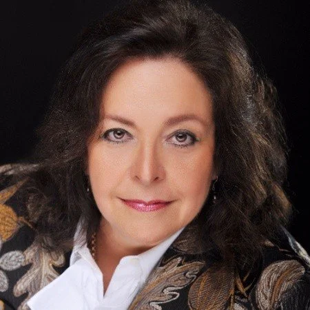 Portrait of a woman with brown hair, wearing a patterned blazer and a white blouse, against a dark background.