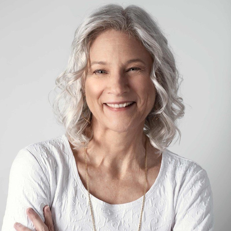 Older woman with gray hair smiling in soft lighting, wearing a white blouse.