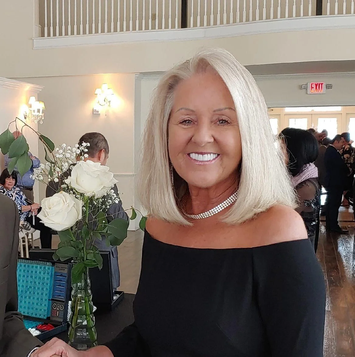 Smiling woman in black dress holding a bouquet of white roses in an indoor event setting.