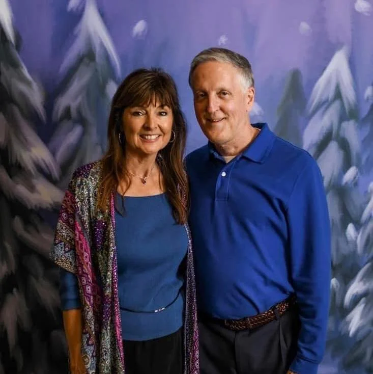 A man and woman smiling, standing in front of a painted winter landscape backdrop with snow-covered trees. The woman is wearing a blue top and patterned shawl, while the man is dressed in a blue polo shirt.
