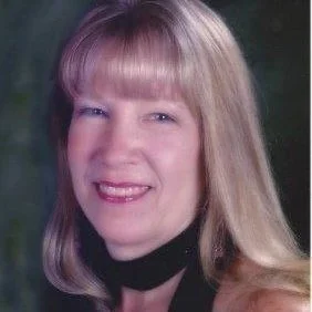 Portrait of a woman with long blonde hair and bangs, wearing a black scarf, smiling against a dark background.