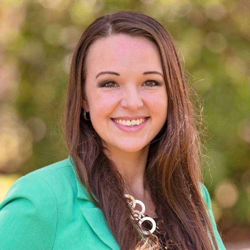 Smiling woman with long brown hair, wearing a green jacket and statement necklace, outdoors with blurred background.