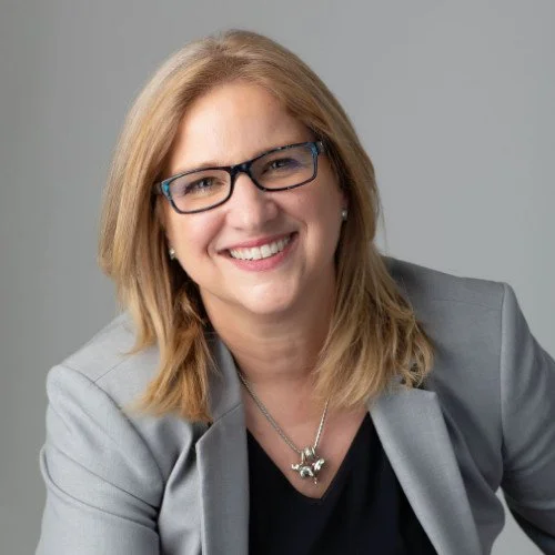 Woman in glasses smiling, wearing a gray blazer and black top