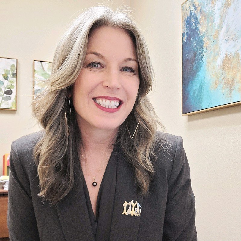 Smiling woman with long hair wearing a black blazer in an office setting.