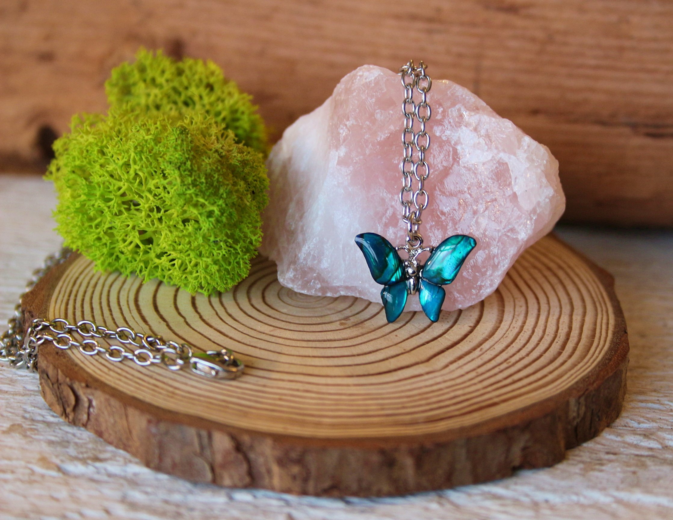 A silver butterfly necklace with blue-green wings draped over a pink salt rock, with green moss and a wooden surface in the background.