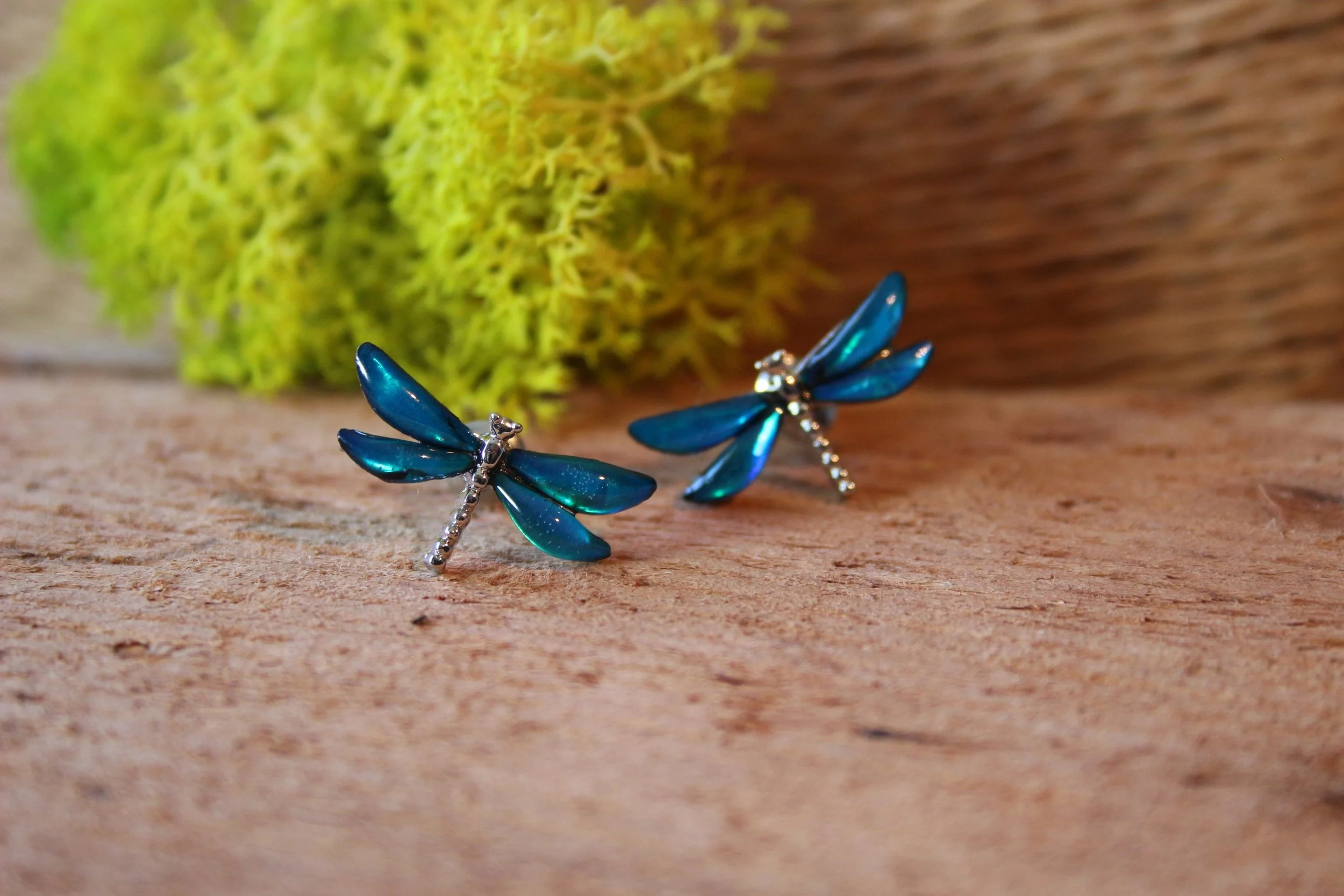 Blue dragonfly earrings on wooden surface with green moss and bark in the background.