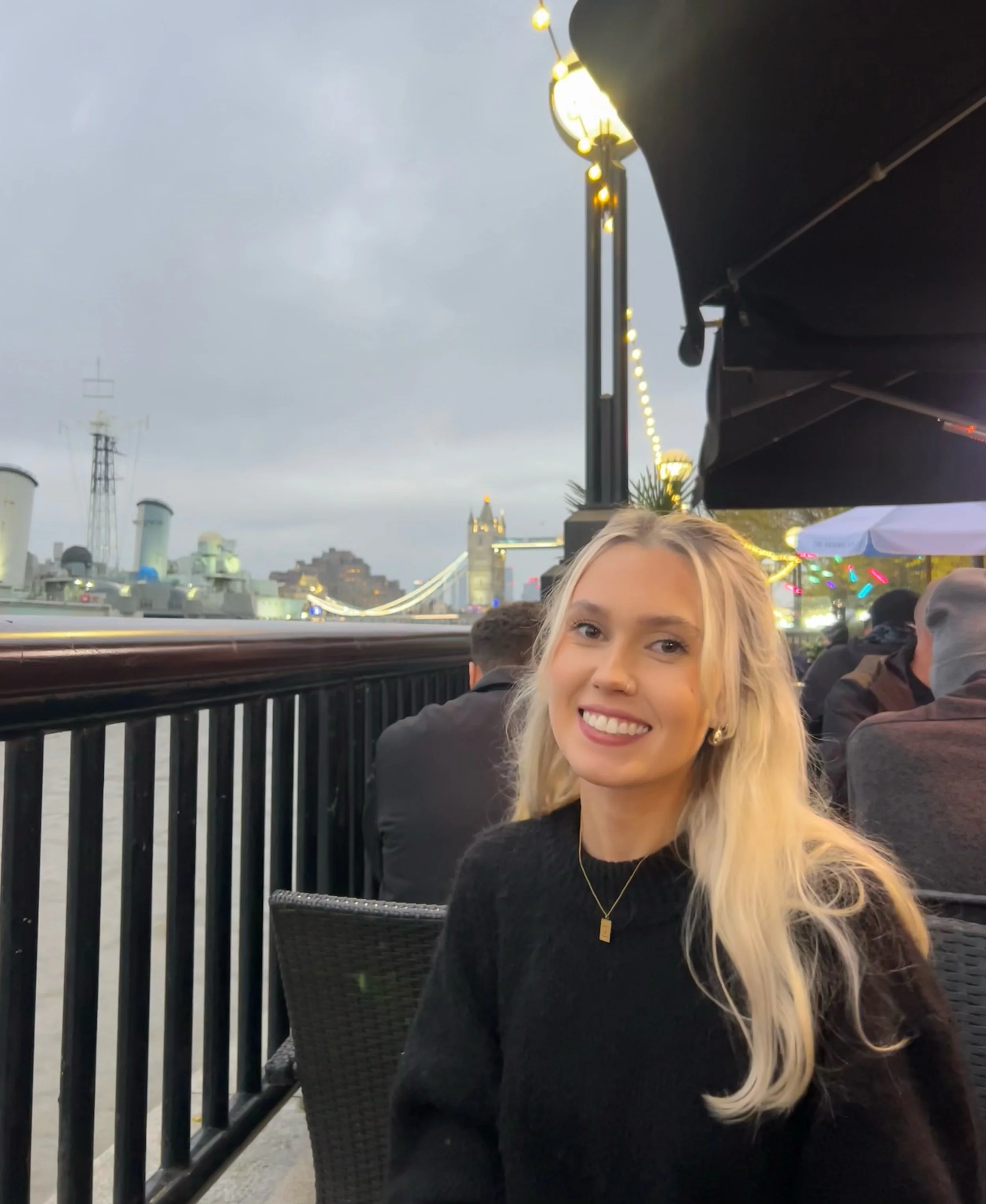 Smiling woman in black sweater sitting outdoors by a riverside, with Tower Bridge in the background, during evening.