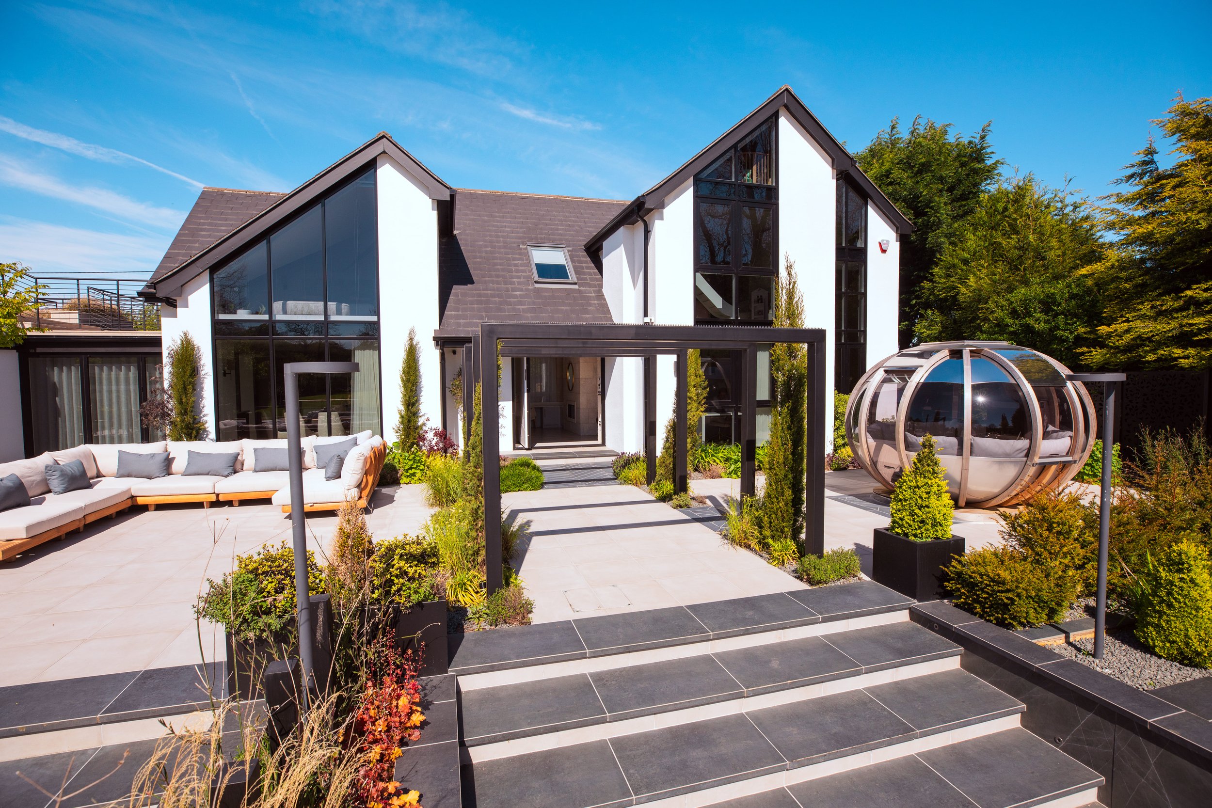 Stunning modern home with a sleek black and white exterior, large floor-to-ceiling windows, and contemporary landscaping. The outdoor space features a luxurious L-shaped sofa, a futuristic glass pod seating area, and a stylish black pergola walkway,