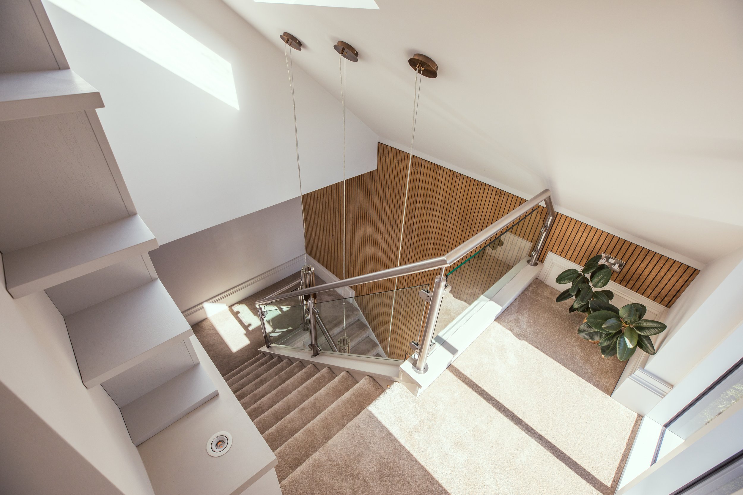 Modern staircase with glass and stainless steel railing, soft beige carpeting, and a slatted wood accent wall. The space is illuminated by natural light from skylights, complemented by elegant pendant lighting. A potted rubber plant adds a touch of g