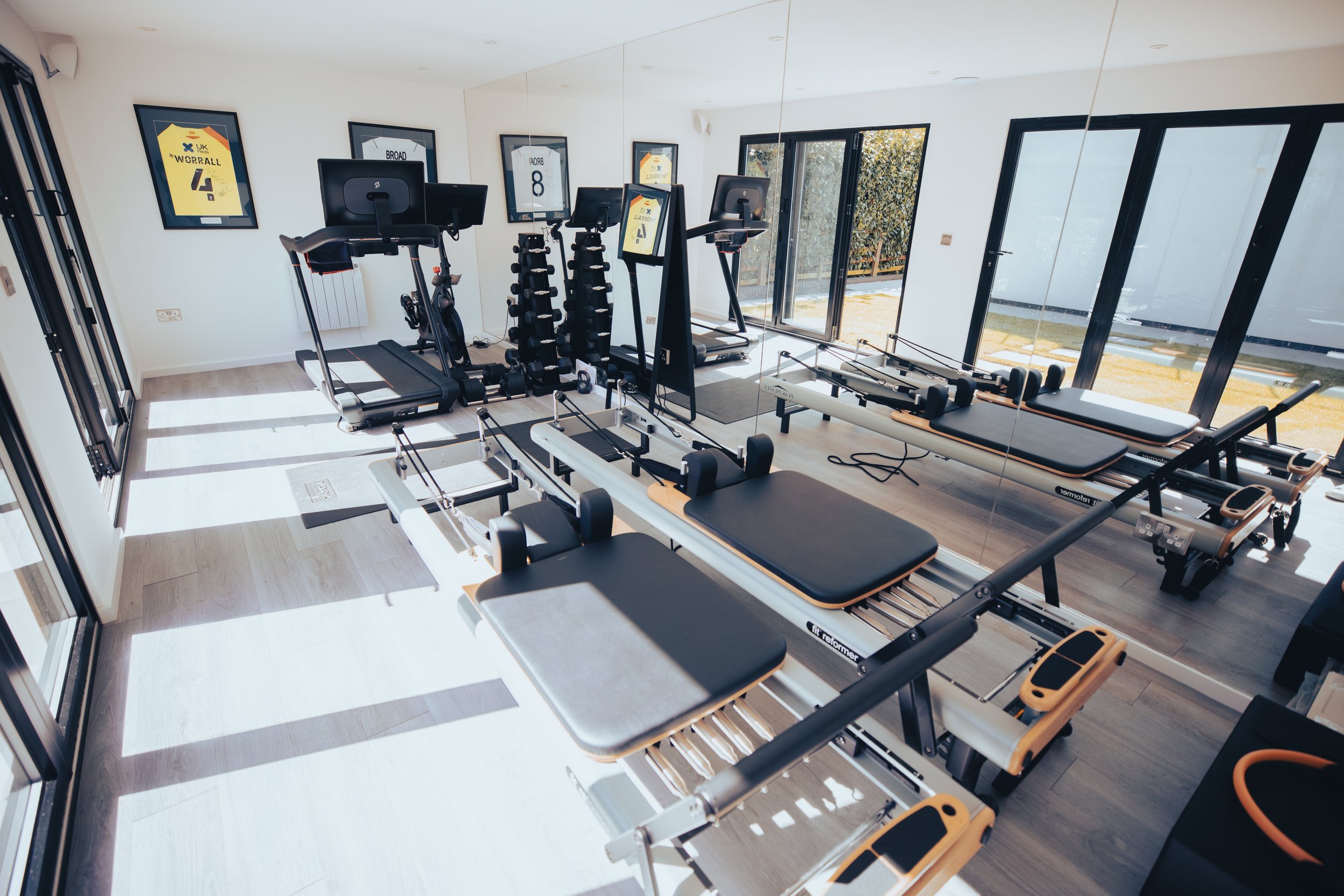Modern home gym with large mirrors, natural light, and high-end fitness equipment, including Pilates reformers, treadmills, free weights, and a stationary bike. The stylish workout space features sleek glass doors opening to an outdoor area, creating