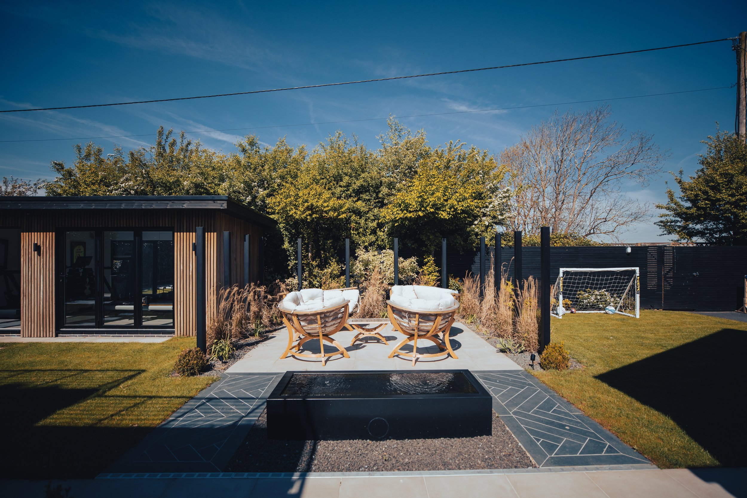 Modern landscaped backyard with a sleek water feature, cozy rattan lounge chairs, and a stylish wooden-clad garden room with large glass doors. The outdoor space features lush greenery, tall ornamental grasses, and a small soccer goal, creating a per
