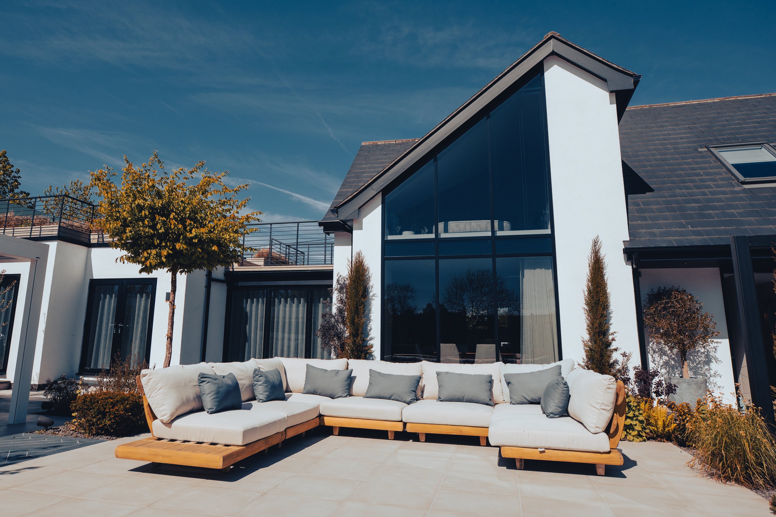Luxury outdoor seating area with a modern L-shaped wooden sofa featuring plush white cushions and grey accent pillows. The space is set against a contemporary white villa with floor-to-ceiling glass windows, a slate roof, and landscaped greenery, cre