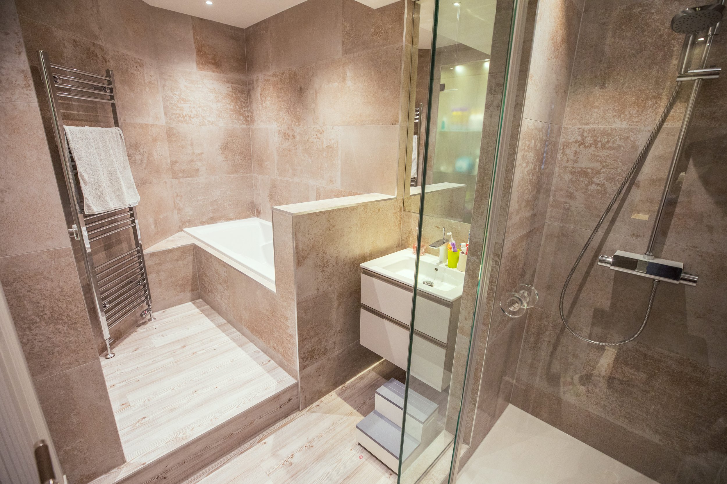 Modern bathroom with a sleek walk-in glass shower, built-in bathtub, and contemporary vanity unit with a white sink. The space features elegant stone-effect wall tiles, a chrome heated towel rail, and light wood flooring, creating a warm and luxuriou