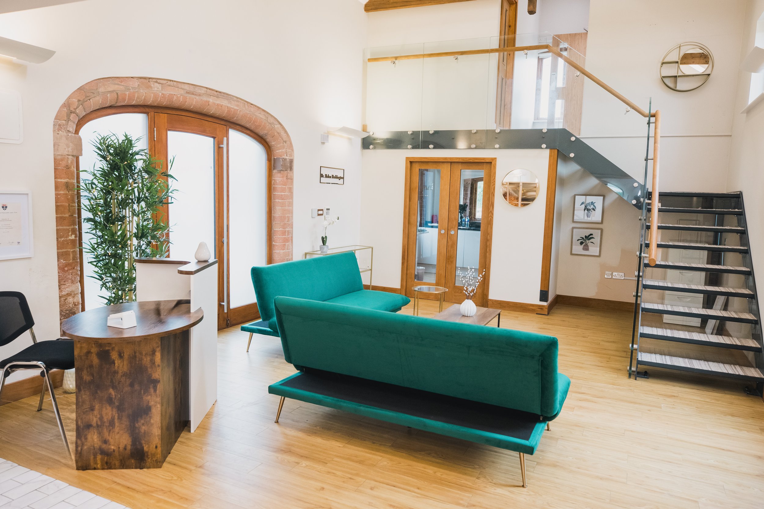 Modern office lounge with stylish emerald green velvet sofas, a sleek glass staircase with wooden accents, and exposed brick archway doors. The space features a mix of contemporary and rustic design elements, including a wooden reception desk, minima