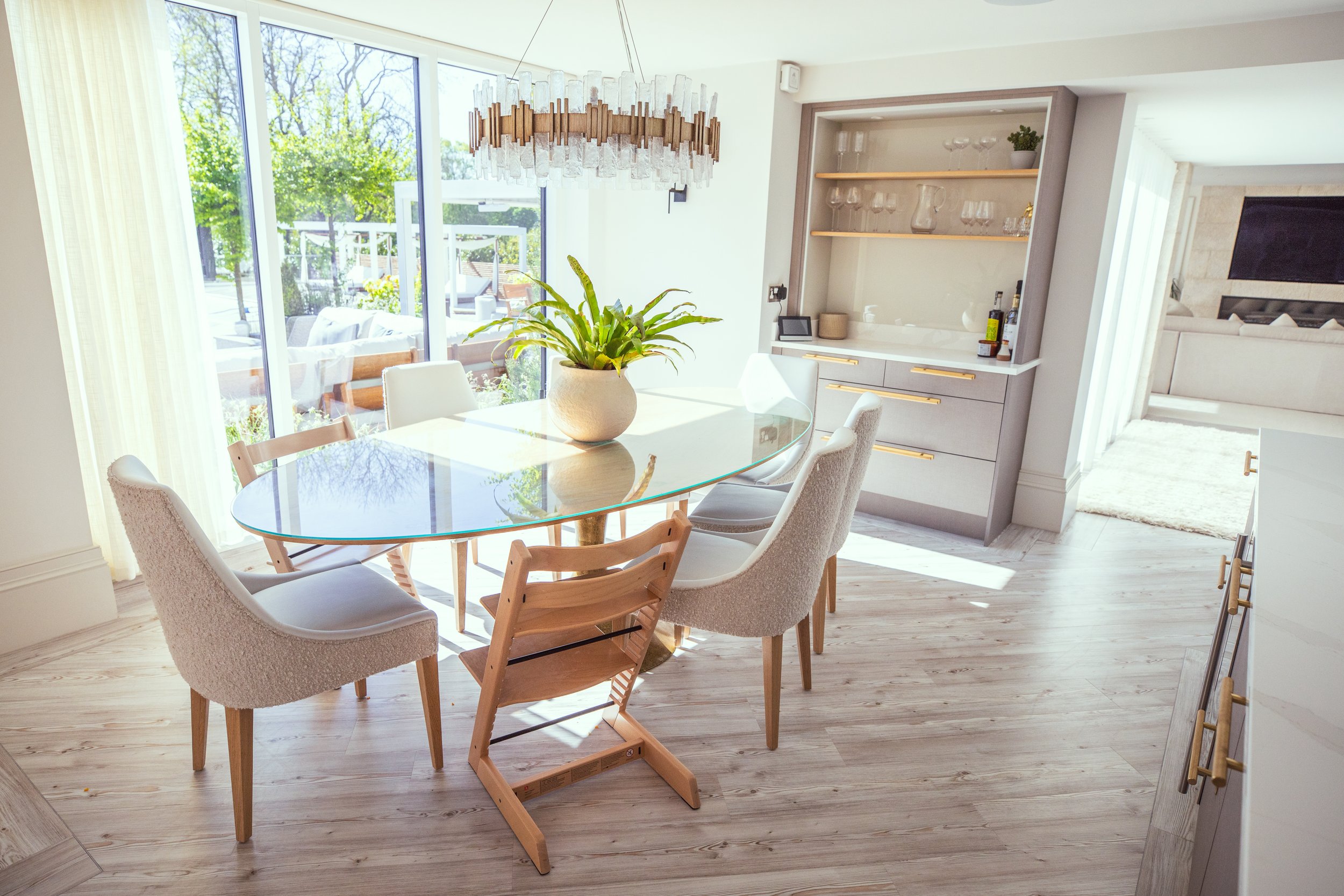 Elegant modern dining area with a round glass table, plush upholstered chairs, and a stylish wooden high chair. A statement chandelier with crystal and gold accents hangs above, while large floor-to-ceiling windows flood the space with natural light.