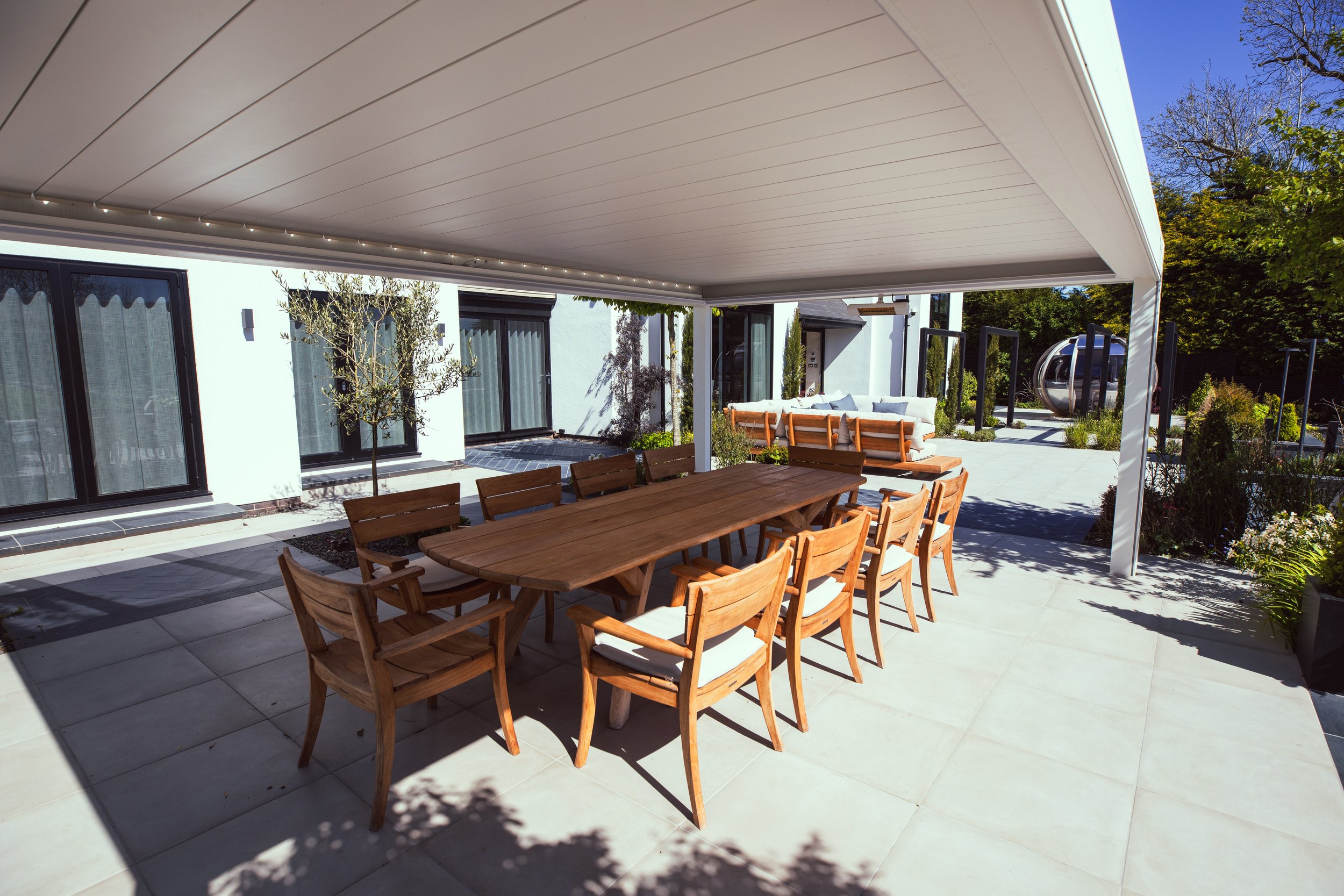 Spacious outdoor dining area with a large wooden table and cushioned chairs under a modern pergola with integrated lighting. The elegant patio features a contemporary white villa, a cozy lounge area with wooden seating, lush landscaping, and a unique