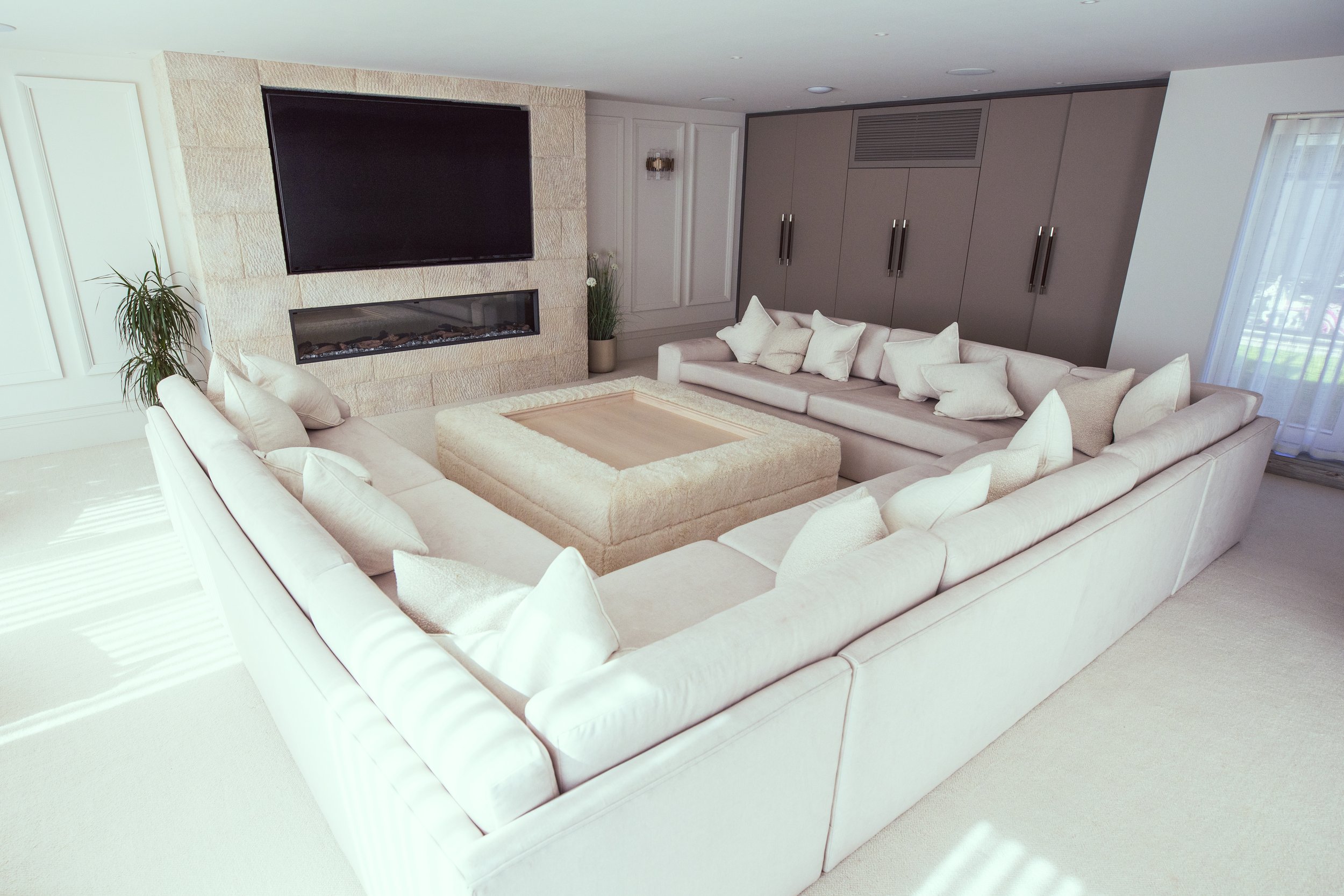 Luxury modern living room with plush cream-colored sectional sofas, soft textured cushions, and a cozy ottoman-style coffee table. The space features a sleek built-in fireplace, a large flat-screen TV, and elegant cabinetry with hidden storage, creat