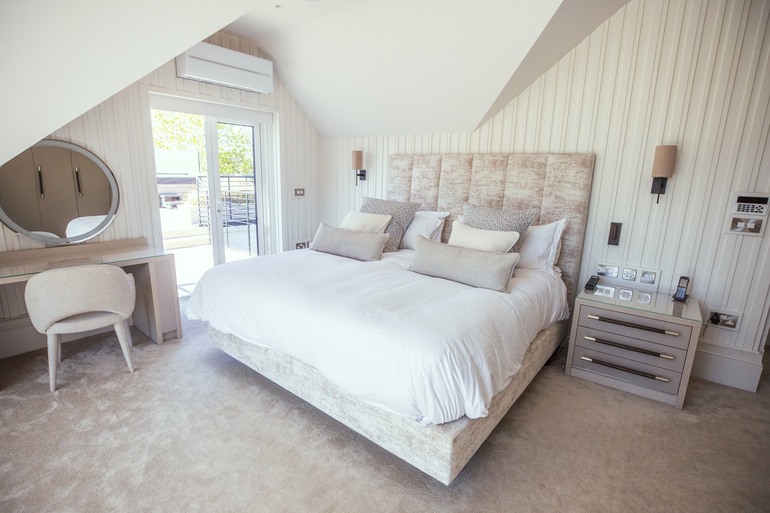 Luxurious modern bedroom with a plush upholstered bed, soft neutral-toned bedding, and an elegant vanity with a round mirror. The space features textured wall paneling, a cozy carpeted floor, and built-in lighting. French doors open to a private balc