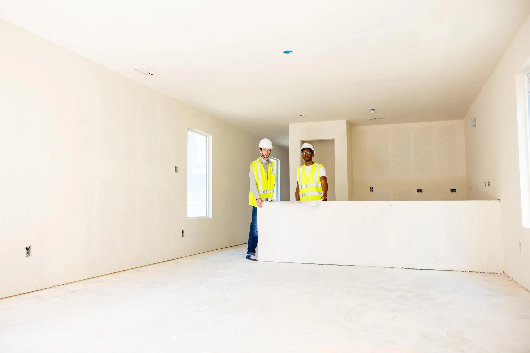 Two construction workers wearing yellow safety vests and white helmets inside an unfinished house with white walls and windows.