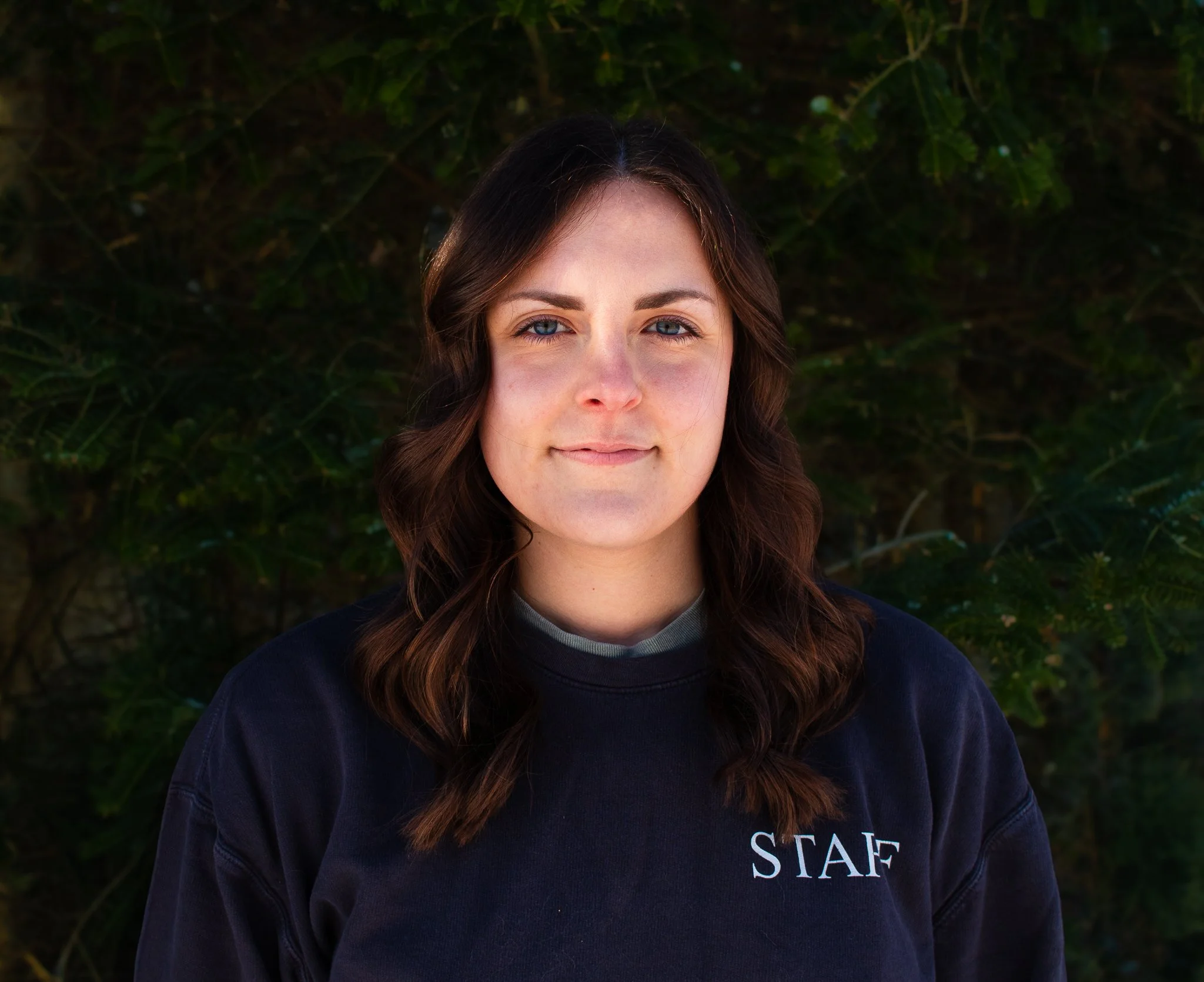 Young woman with wavy brown hair and blue eyes standing outdoors against a background of green foliage, wearing a black shirt with the word 'STAFF' on it.