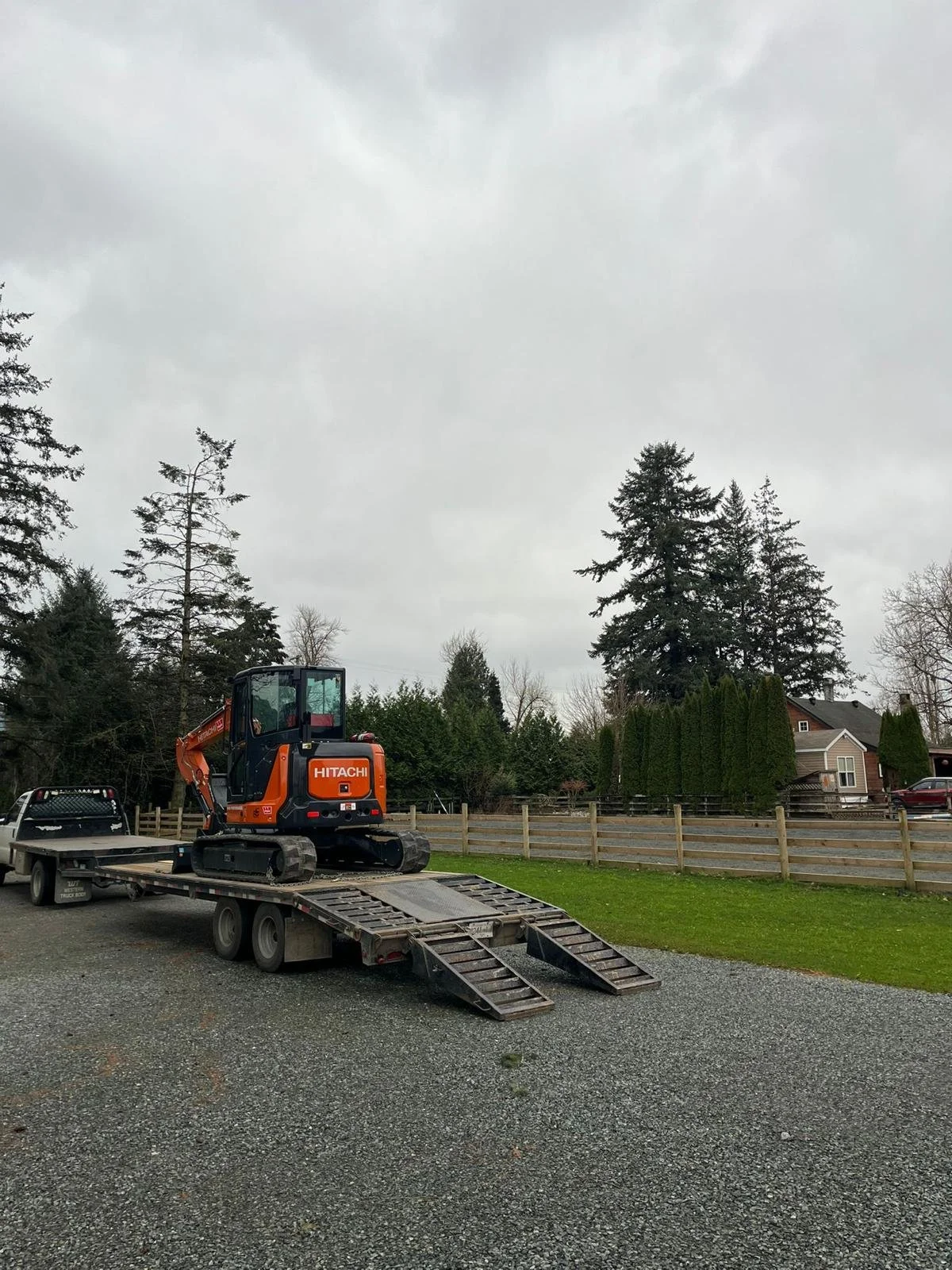 Orange Hitachi mini excavator on a trailer hitched to a pickup truck in a rural setting with trees and a house in the background.