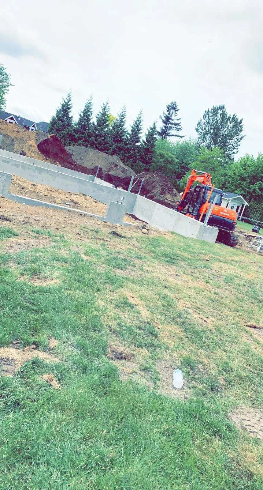 Construction site with an orange excavator near a concrete foundation, surrounded by dirt and grass, and trees in the background.