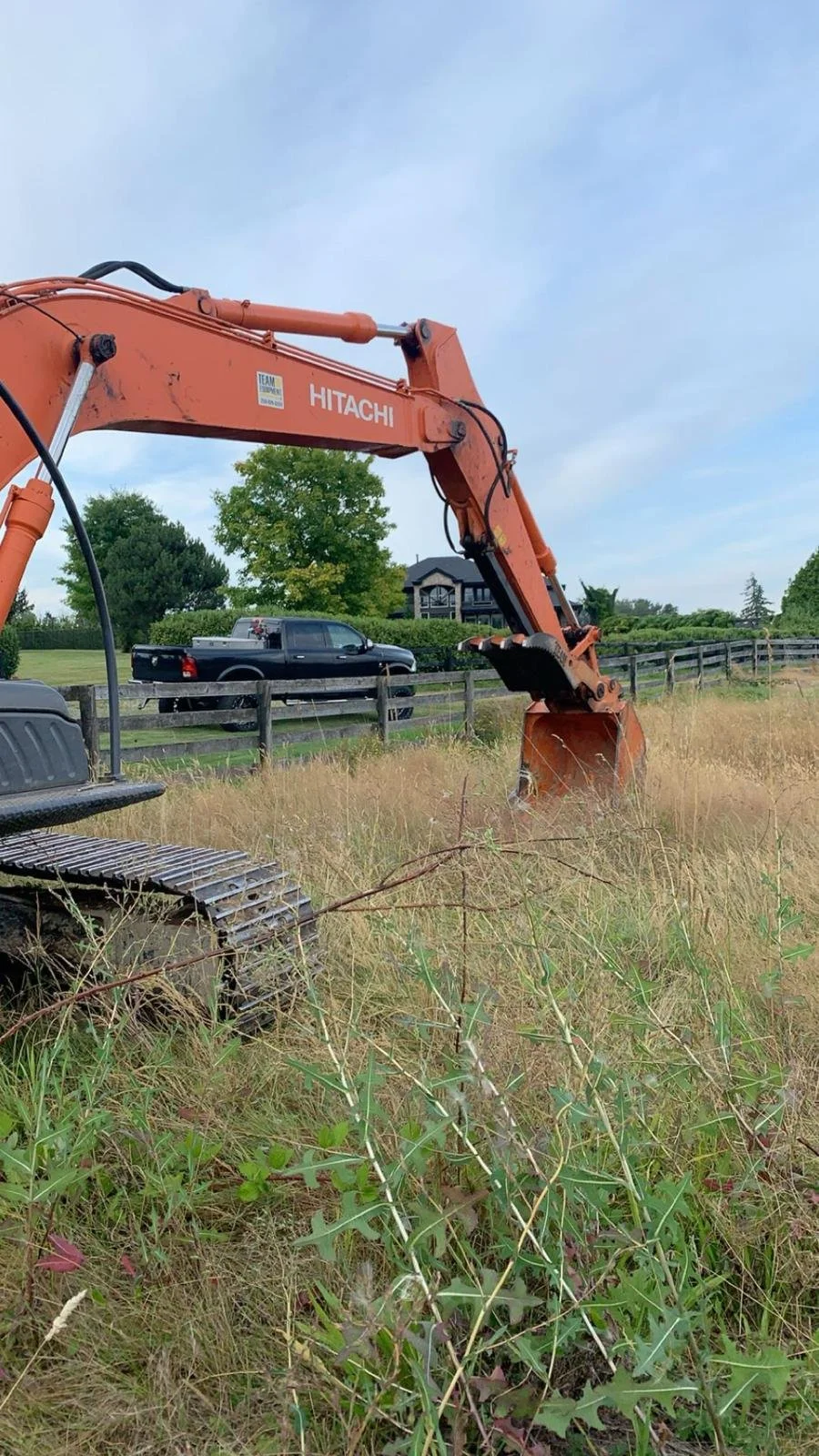 Orange excavator in a grassy field with a black truck and house in the background.