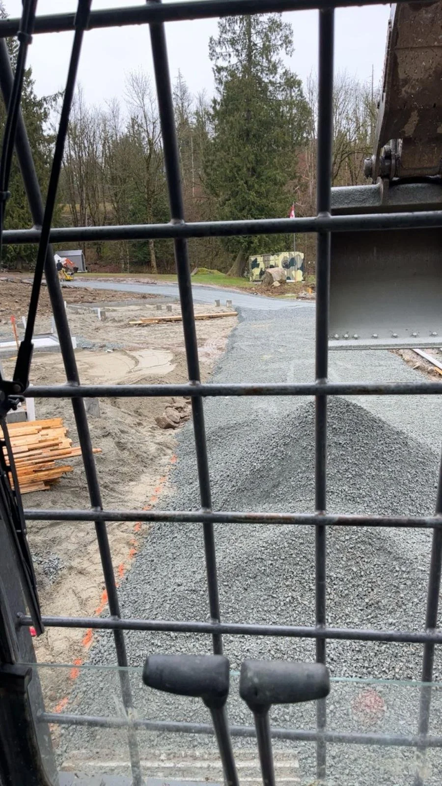 View from inside construction vehicle cabin showing gravel road, piles of construction materials, trees, and overcast sky.