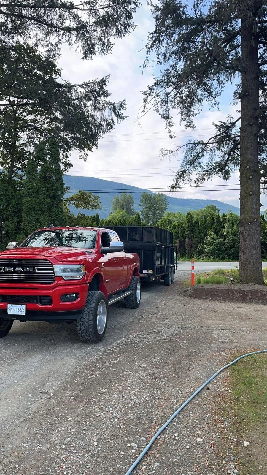 Red pickup truck towing a large black trailer on a gravel driveway, surrounded by trees and mountains in the background.