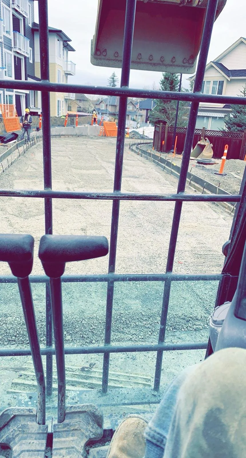 View from inside a construction vehicle, overlooking a construction site with gravel, buildings, and workers in hard hats.