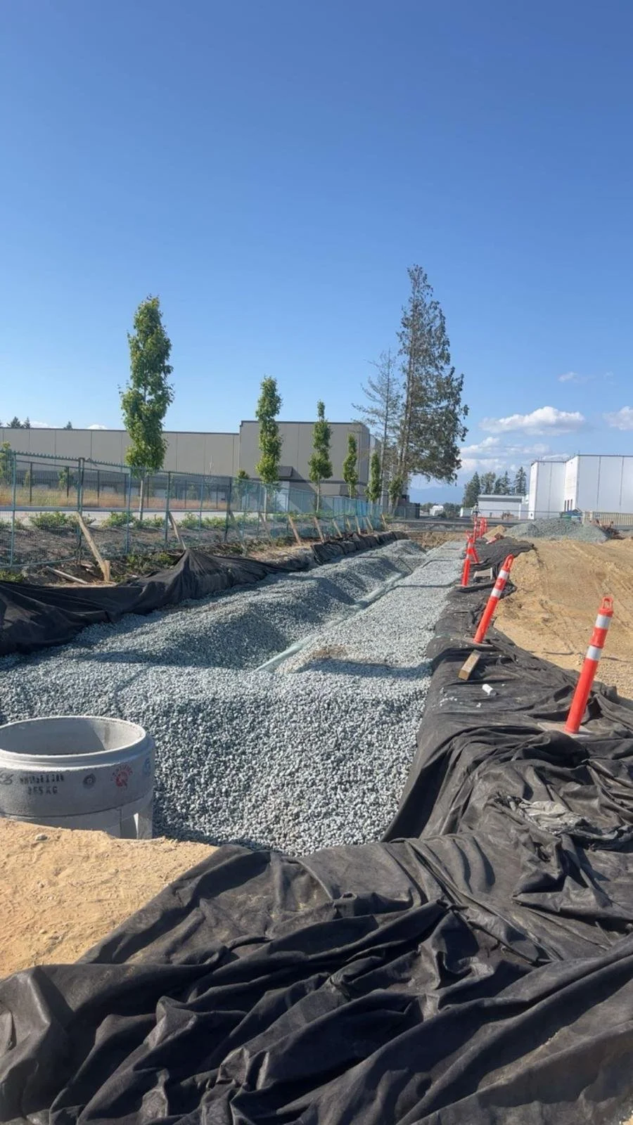Construction site with gravel path, orange cones, and trees; building in the background.